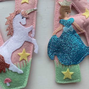K&ouml;nnte beinhalten: Eine rosa Filzbuchstaben-Dekoration mit einem wei&szlig;en Einhorn mit kupferfarbener Glitzerm&auml;hne und Horn sowie einer Figur in einem blauen Glitzerkleid mit goldener Krone. Das Design umfasst gelbe Filzsterne, gr&uuml;nes Filzgras und einen kleinen rosa Pilz.