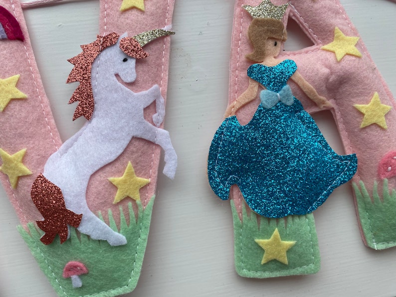 K&ouml;nnte beinhalten: Eine rosa Filzbuchstaben-Dekoration mit einem wei&szlig;en Einhorn mit kupferfarbener Glitzerm&auml;hne und Horn sowie einer Figur in einem blauen Glitzerkleid mit goldener Krone. Das Design umfasst gelbe Filzsterne, gr&uuml;nes Filzgras und einen kleinen rosa Pilz.