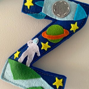 May include: A dark blue felt letter Z decoration featuring space-themed appliqu&eacute;s including a light blue rocket ship with a gray porthole, a green planet with an orange ring, a white astronaut figure, and several small yellow felt stars.