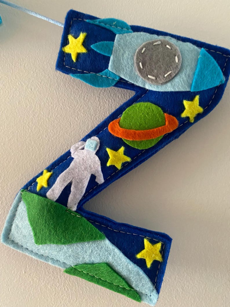 May include: A dark blue felt letter Z decoration featuring space-themed appliqu&eacute;s including a light blue rocket ship with a gray porthole, a green planet with an orange ring, a white astronaut figure, and several small yellow felt stars.