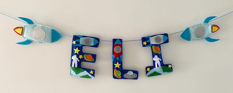 May include: A felt banner spelling the name ELI in blue letters, decorated with space-themed appliqu&eacute;s including rockets, planets, stars, astronauts, and a flying saucer. The banner is strung on a thin cord with two additional rockets at the ends.