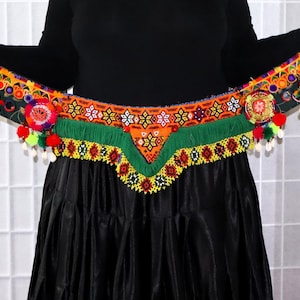 May include: A colourful beaded belt with fringe and tassels. The belt is made of various colours, including red, orange, yellow, green, blue, and white. The belt is worn around the waist of a person wearing a black skirt.