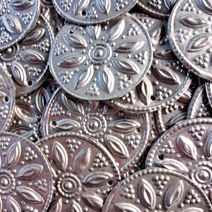 Könnte beinhalten: Nahaufnahme einer Sammlung silberfarbener Metallverzierungen. Jedes kreisförmige Stück weist ein erhabenes Blumendesign mit einem Mittelkreis und blütenblattartigen Formen sowie kleine Löcher zum Befestigen auf. Die Teile sind aufeinander gestapelt.