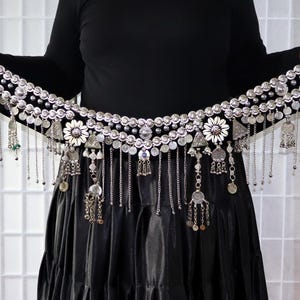 Könnte beinhalten: Ein silberner Bauchtanzgürtel mit baumelnden Ketten und dekorativen Münzen. Der Gürtel ist mit weißen, blumenförmigen Akzenten verziert und wird über einem schwarzen Kleid getragen.