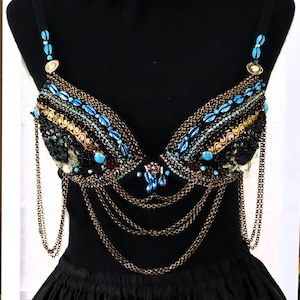 Può includere: Un reggiseno nero con perline dorate e blu. Il reggiseno presenta intricati dettagli a catena ed è adornato con perline color turchese. Anche le spalline sono impreziosite da perline. Il reggiseno è esposto su un manichino nero.