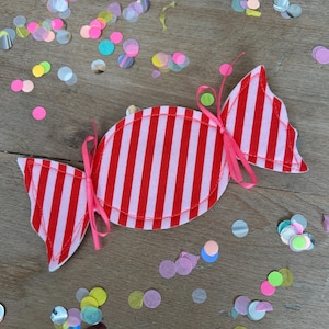 Könnte beinhalten: Eine stoffliche Süßigkeiten-Dekoration mit rot-weißen Streifen, gebunden mit rosa Bändern. Bunte Konfetti in verschiedenen Formen sind um das Objekt herum verstreut, was eine festliche und spielerische Szene erzeugt.