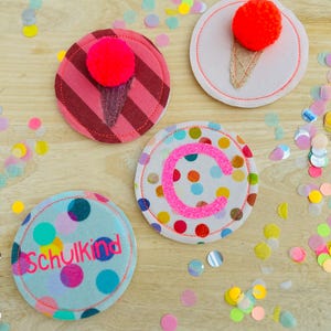Könnte beinhalten: Vier bunte, runde Filzuntersetzer mit dekorativen Elementen. Zwei Untersetzer zeigen ein Eiscreme-Design mit rotem Pompon. Ein Untersetzer hat den rosa Buchstaben "C", und ein anderer sagt "Schulkind" in Rosa. Konfetti ist verstreut.