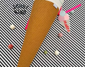 Pode incluir: Um recipiente de tecido em forma de cone de waffle marrom com uma parte superior branca e uma fita rosa. O recipiente está cheio de um coelho de tecido branco com orelhas rosa e um tecido listrado rosa e branco. Uma etiqueta rosa com o texto "Madita" e uma imagem de três bolas de sorvete está presa ao recipiente. O recipiente está sobre um fundo listrado preto e branco.