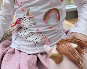 May include: A white long-sleeved shirt with the name "Carlota" spelled out in colourful fabric letters. The shirt features a rainbow and a unicorn with black polka dots.