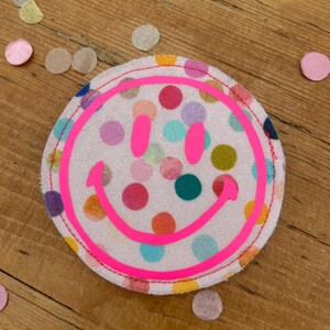 Puede incluir: Un parche de tela rosa con una carita sonriente rosa brillante y un patr&oacute;n de lunares de colores.