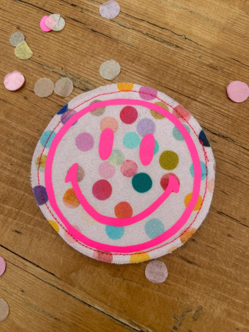 Puede incluir: Un parche de tela rosa con una carita sonriente rosa brillante y un patr&oacute;n de lunares de colores.