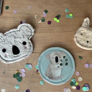Könnte beinhalten: Eine Sammlung von Artikeln zum Thema Koala, darunter ein Plüsch-Koala-Gesicht, ein runder Untersetzer mit einer Koala-Illustration und ein Plüsch-Hasen-Gesicht. Die Artikel sind hellgrau und weiß, mit schwarzen und rosa Akzenten. Bunte Konfetti sind verstreut.