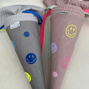 Könnte beinhalten: Zwei graue Stoffkegel mit aufgedruckten Smiley-Gesichtern. Ein Kegel hat ein blaues Band und den aufgedruckten Text "Mila Louise". Der andere Kegel hat ein rosa Band und ein hellgrünes Band.
