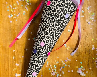 Könnte beinhalten: Eine kegelförmige Schultüte mit einem schwarz-weißen Leopardenmuster. Die Tüte ist mit rosa, lila und weißen Punkten verziert und hat rosa und rote Bänder. Eine blaue Tüllschleife schmückt die Oberseite.