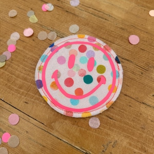 Könnte beinhalten: Ein rosa Stoff-Patch mit weißem Hintergrund und einem bunten Punkte-Muster. Der Patch zeigt ein rosa Smiley-Gesicht mit gelbem Rand.