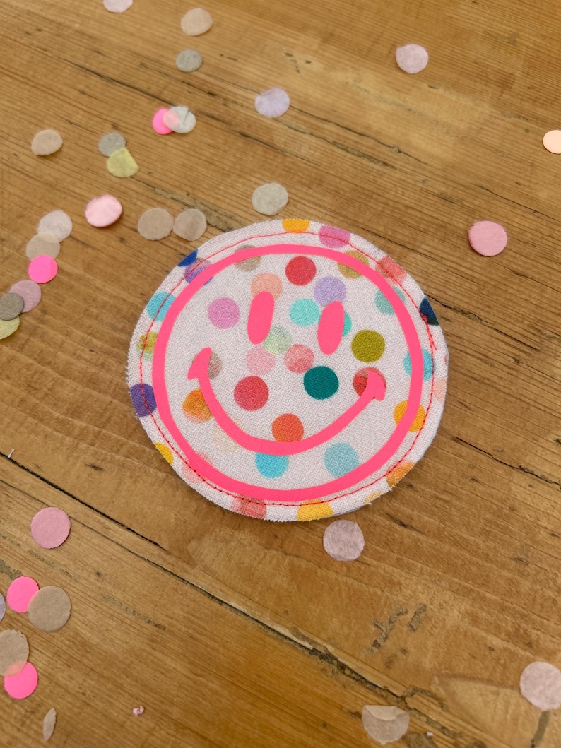 Puede incluir: Un parche de tela rosa con fondo blanco y un patr&oacute;n de lunares multicolores. El parche presenta una cara sonriente rosa con un contorno amarillo.