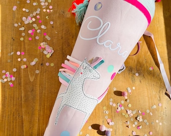 May include: A pink and blue school cone with a unicorn design and the name "Clara" written on it. The cone is surrounded by confetti.