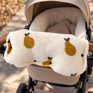 Könnte beinhalten: Ein cremefarbener, strukturierter Kinderwagen-Lenkerbezug aus Sherpa-Stoff mit einem wiederkehrenden Muster aus senfgelben Birnen mit kleinen schwarzen Stielen. Der Bezug ist um den braunen Lederimitat-Lenker eines beigen Kinderwagens gewickelt.