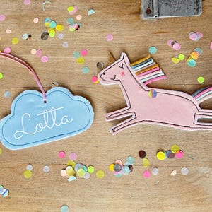 Könnte beinhalten: Ein hellblaues Wolken-Tag mit dem Namen "Lotta" in weißer Schrift, neben einem rosa Einhorn mit Regenbogenmähne und -schwanz. Bunte Konfetti sind auf einer Holzoberfläche verstreut.