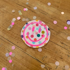 Puede incluir: Un parche de tela sonriente rosa con fondo blanco y lunares de colores. El parche est&aacute; sobre una superficie de madera con confeti esparcido a su alrededor.