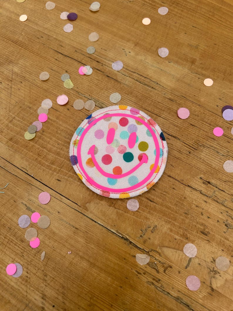 Puede incluir: Un parche de tela sonriente rosa con fondo blanco y lunares de colores. El parche est&aacute; sobre una superficie de madera con confeti esparcido a su alrededor.