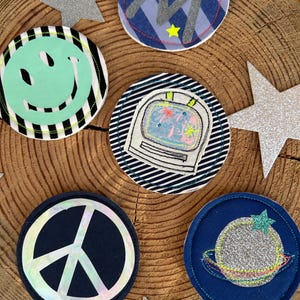 Könnte beinhalten: Eine Sammlung bunter, runder Stoffaufnäher mit verschiedenen Designs, darunter ein Smiley, ein Retro-Computer, ein Friedenszeichen und ein Planet. Silberne Glitzersterne sind um die Aufnäher herum verstreut. Die Aufnäher liegen auf einer Holzoberfläche.