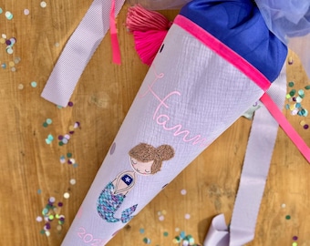 May include: A purple and pink school cone with a mermaid design and the name "Fami" embroidered on it. The cone has a pink ribbon and a blue bow with a pink tassel.