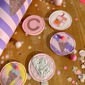 Könnte beinhalten: Fünf bunte Filzpatches mit verschiedenen Designs, darunter ein Eiswaffel, eine Wolke, ein Saftkarton und der Buchstabe C. Die Patches befinden sich auf einer Holzoberfläche mit rosa und weißen Konfetti.