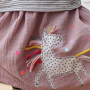 Può includere: Una gonna rosa con un'applicazione di unicorno. L'unicorno è bianco con pois neri e ha una criniera e una coda arcobaleno. La gonna è decorata con punti colorati.