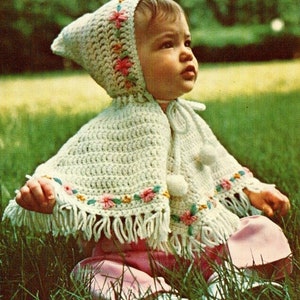 Puede incluir: Un poncho de bebé blanco de ganchillo con capucha y ribete floral. El poncho tiene flecos alrededor del borde inferior y un pompón en la capucha.