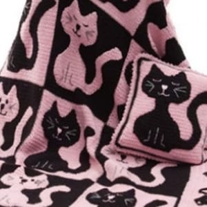May include: A pink and black crocheted throw blanket with a cat design. The blanket features a black cat pillow with a pink background.