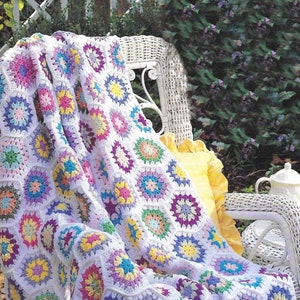 Op de afbeelding: Een wit gehaakt deken met kleurrijke vierkanten, met verschillende tinten roze, paars, blauw, geel en groen. Het deken is gedrapeerd over een witte rieten stoel met een geel kussen.