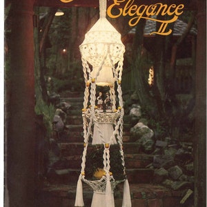 Könnte beinhalten: Ein weißer Makramee-Pflanzenhänger mit drei Ebenen, mit einem weißen Makramee-Lampenschirm oben, gelben Perlen und weißen Quasten. Der Hänger hängt in einer Gartenumgebung mit einem Ziegelweg und grünem Laub. Der Text "Macrame Elegance II" ist oben im Bild sichtbar.