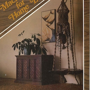 Puede incluir: Un armario de madera marrón con dos puertas se encuentra en una habitación con un macramé colgante para plantas. El macramé está hecho de cuerda natural y tiene un estante de madera. La habitación tiene una alfombra marrón y un cuadro en la pared.