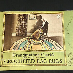 Op de afbeelding: Een vintage boekomslag met een groene achtergrond en de titel "Grandmother Clark's How to Make Hand Made Crocheted Rag Rugs" in grote, vetgedrukte letters. De omslag toont een illustratie van een vrouw die een tapijt haakt in een kamer met een raam en een klok.