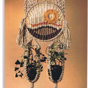 May include: A macrame plant hanger with two pots and plants. The hanger is made of natural colored cord and has a decorative design with a brown and white center and a yellow and orange flower. The hanger is hanging from a ceiling hook.