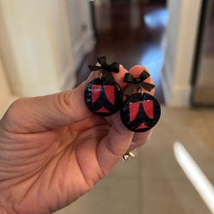 Könnte beinhalten: Runde, schwarz-rote Ohrringe mit Schleife. Die Ohrringe haben einen schwarzen Hintergrund mit einem roten Design. Jeder Ohrring hat eine kleine schwarze Schleife oben. Die Ohrringe werden von einer Hand gehalten.