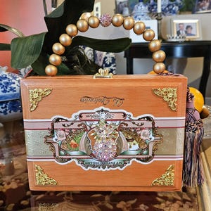 May include: A handcrafted wooden handbag made from a cigar box. The box is light brown with gold accents and a decorative label. The handle is made of gold-colored beads, with a pink jeweled center.