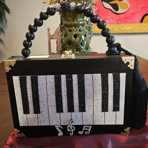 May include: Black rectangular handbag with a piano key design on the front. The bag has a beaded handle, gold-colored corner accents, and a black tassel. Musical note embellishments are below the piano keys.