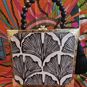 May include: A square box-shaped handbag featuring a black and white ginkgo leaf pattern. The bag has gold-tone metal corner protectors, a gold clasp, and a handle made of large black beads with a central rhinestone bead. A black tassel hangs from the side.