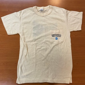 May include: A white t-shirt with a pocket on the left side. The pocket has the text "UNIVERSAL RECTIFIER" and a blue logo with a stylized "U" inside a square.