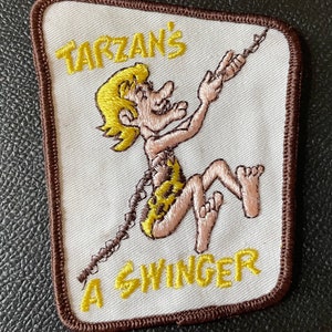 May include: A brown and white embroidered patch with a cartoon image of Tarzan swinging on a vine. The text "Tarzan's A Swinger" is embroidered in yellow.
