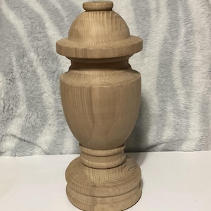 May include: A light-colored, unfinished wooden urn with a domed lid and a decorative base. The urn has a classic shape, with a wide body and a narrow neck. The wood grain is visible, giving it a natural look. The urn is suitable for home decor.