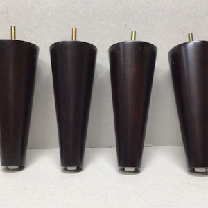 May include: Four dark brown wooden furniture legs with a tapered design and a metal screw at the top. Each leg has a white plastic foot pad.