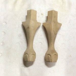 May include: Two unfinished wooden furniture legs, each featuring a square top, a flared midsection, and a carved foot design. The legs are light brown and sit on a white surface.