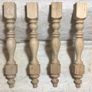 May include: Four unfinished wooden table legs. Each leg features a square top, a turned central column with decorative details, and a base with a rounded foot. The legs are made of light-colored wood.