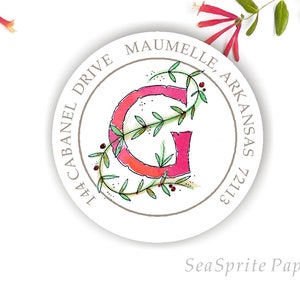 May include: A round, white sticker with a gray border and the text "1444 CABANEL DRIVE MAUMELLE, ARKANSAS 72113". The center of the sticker features a pink letter "G" surrounded by green leaves and red berries.