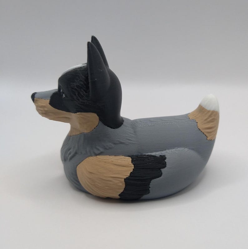 May include: A gray, black, and brown rubber duck with a blue heeler dog design. The duck has a white tip on its tail.