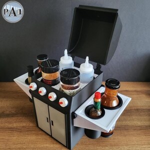 Puede incluir: Un organizador en miniatura con forma de parrilla negra y gris con tapa abatible. Contiene varios frascos de especias, botellas exprimibles y botellas de condimentos. El organizador tiene bandejas laterales blancas y perillas de control rojas y blancas.