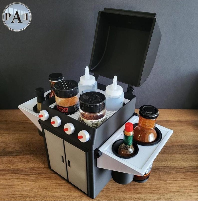 Puede incluir: Un organizador en miniatura con forma de parrilla negra y gris con tapa abatible. Contiene varios frascos de especias, botellas exprimibles y botellas de condimentos. El organizador tiene bandejas laterales blancas y perillas de control rojas y blancas.